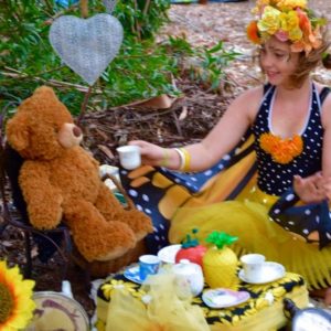 A woman dressed as a butterfly is sitting next to a teddy bear.