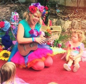 A woman dressed in a fairy costume is playing an ukulele in front of a group of children.