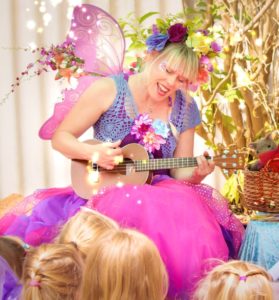 A woman in a fairy costume playing an ukulele in front of a group of children.