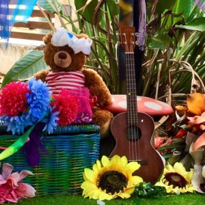 A teddy bear sits next to a basket of flowers and a ukulele.