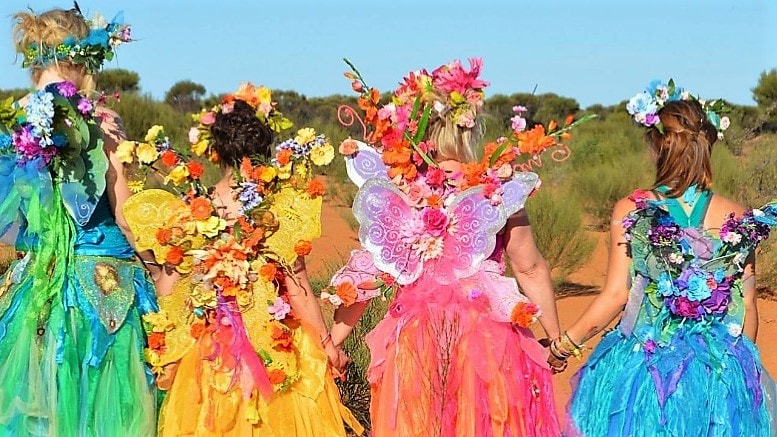 A group of people wearing colorful, elaborate costumes adorned with flowers and wings, walking outdoors, viewed from behind.