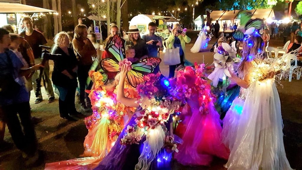 People in colorful, illuminated costumes at a night festival, including butterfly wings and floral dresses, among a crowd.