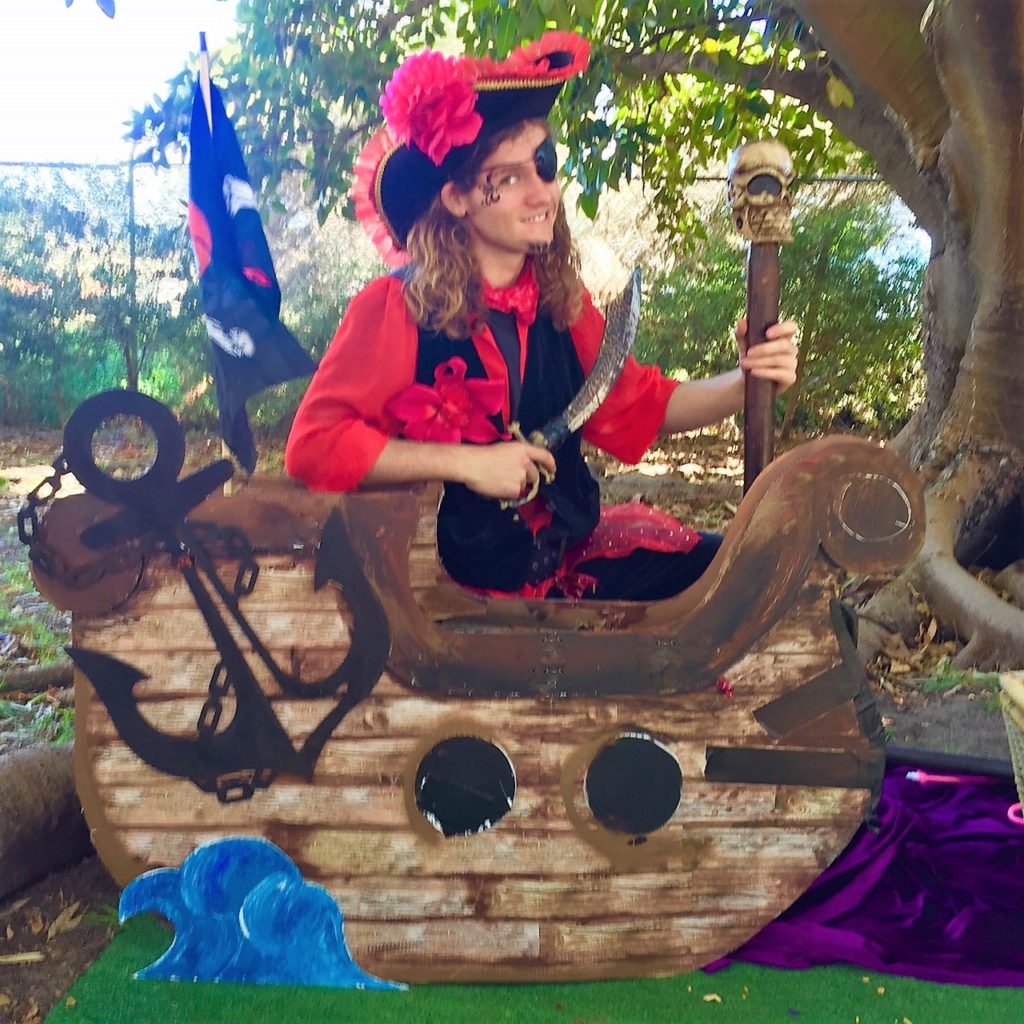 Man in pirate costume sitting in a small wooden ship prop, holding a staff, with pirate flags and greenery in the background.