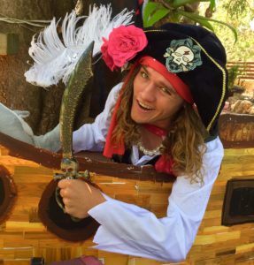 A person dressed as a pirate, smiling and holding a curved sword, standing in front of a wooden structure with feathery decorations.