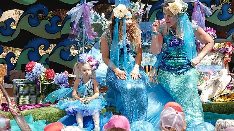 Two performers dressed as mermaids in sequined, blue costumes talking to a child, surrounded by colorful decorations on a parade float.